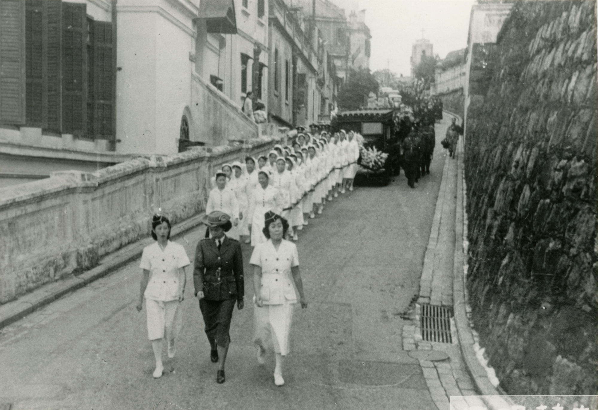 Nurses of the St. John Ambulance Brigade and the hearse move along Seymour Road. In 1915, Ho Kom-tong initiated the founding of the St. John Ambulance Brigade in Hong Kong. With funds donated by Ho, the Brigade was established in the following year.
